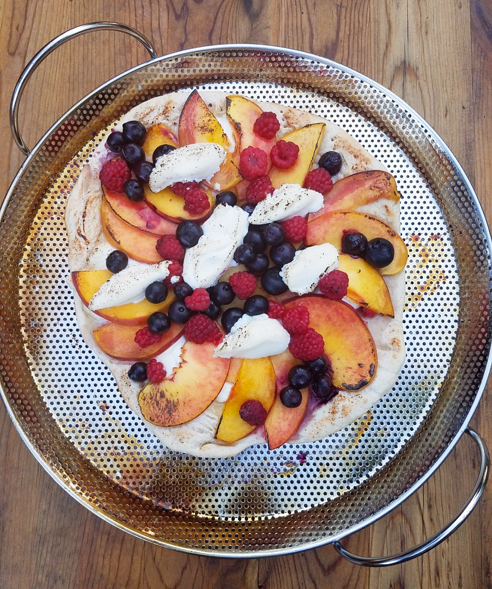 An RSVP Endurance Precision Pierced Pizza Pan sits on a wooden table. The pan has a fruit pizza on it.