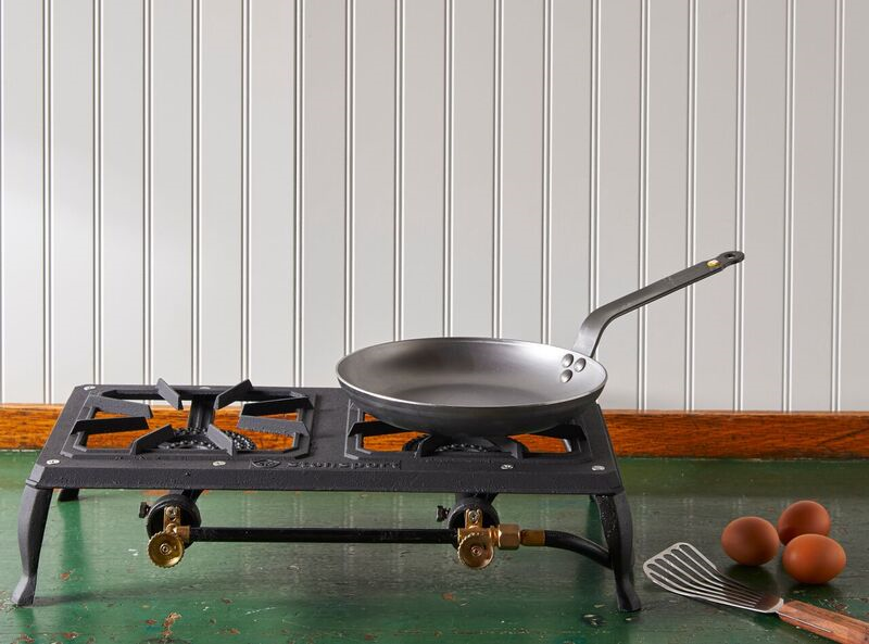 A DeBuyer Mineral B Carbon Steel Omelette Pan rests on a portable stovetop, which sits on a green table in front of a white wall.