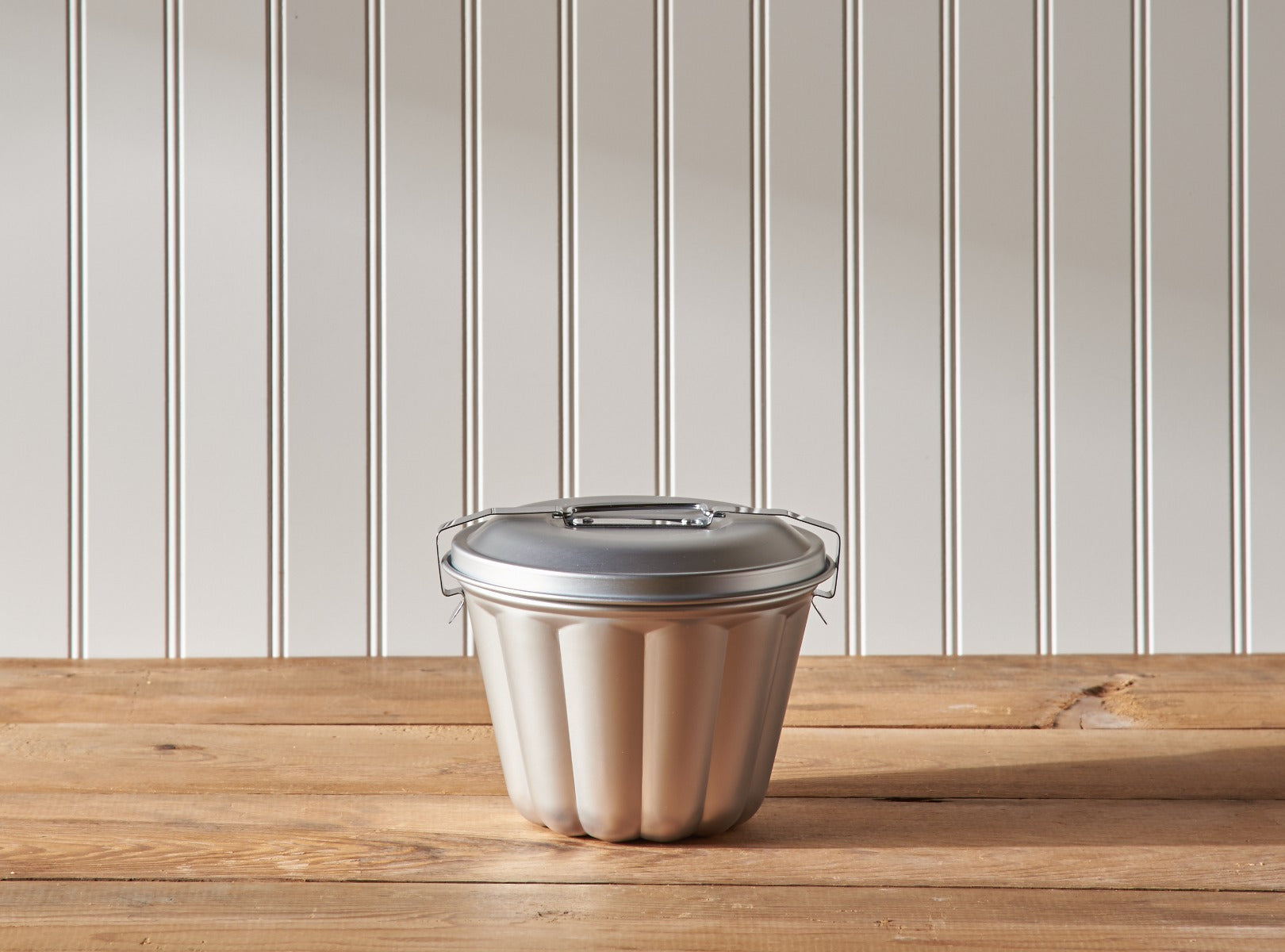 Stainless steel lid on beige food container on wooden table against striped wall.