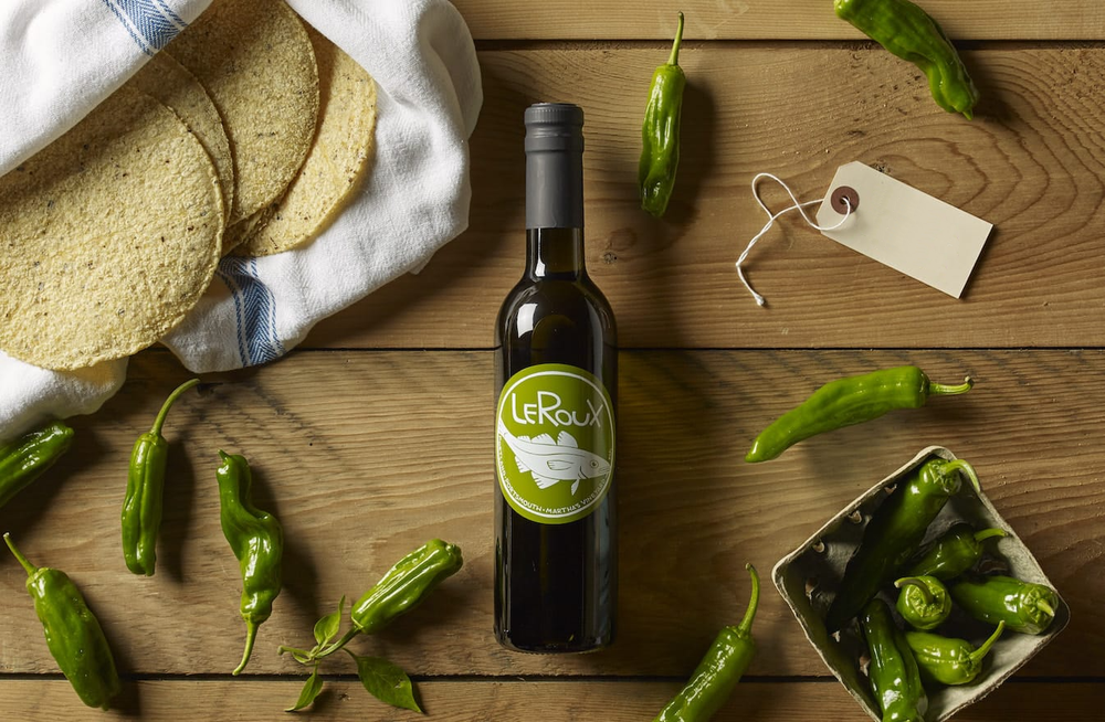 Olive oil bottle on wooden table with green chilies and tortillas.