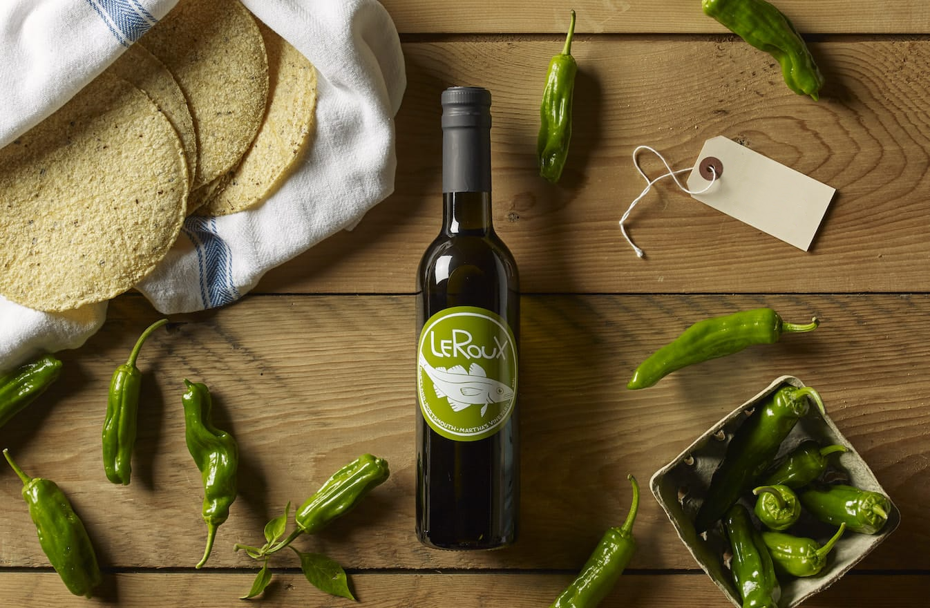 Olive oil bottle on wooden table with green chilies and tortillas.