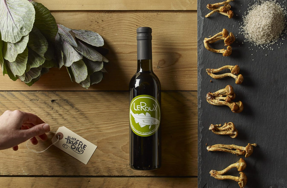 Bottle of LeRoux black truffle oil with mushrooms and sea salt on wooden table.