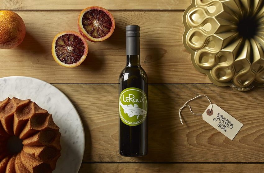 Stylish bottle of Leroux olive oil, blood oranges, and bundt cake pan on wooden table.