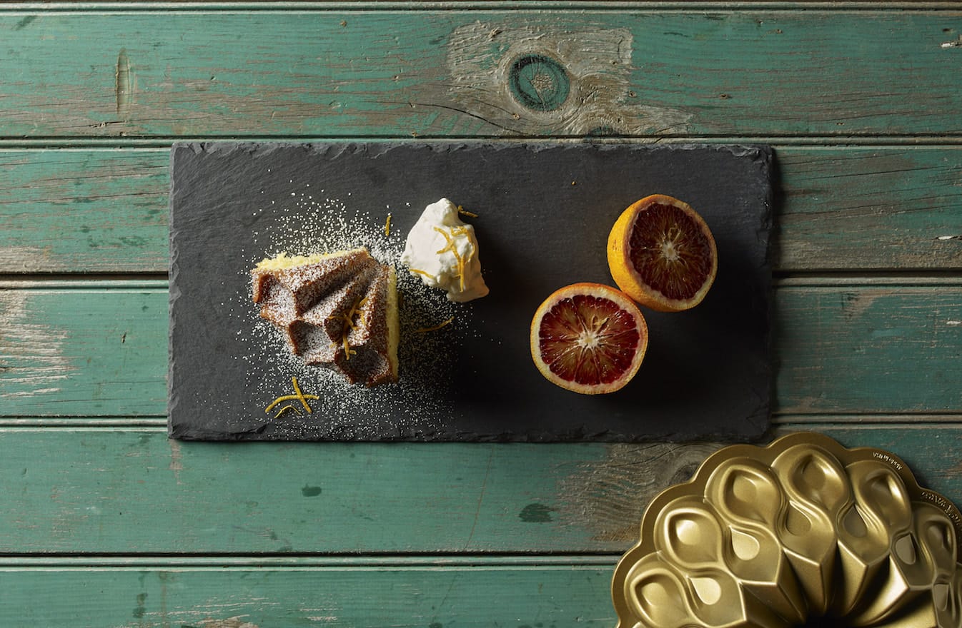 Slice of lemon cake with powdered sugar, cream, and blood oranges on slate board