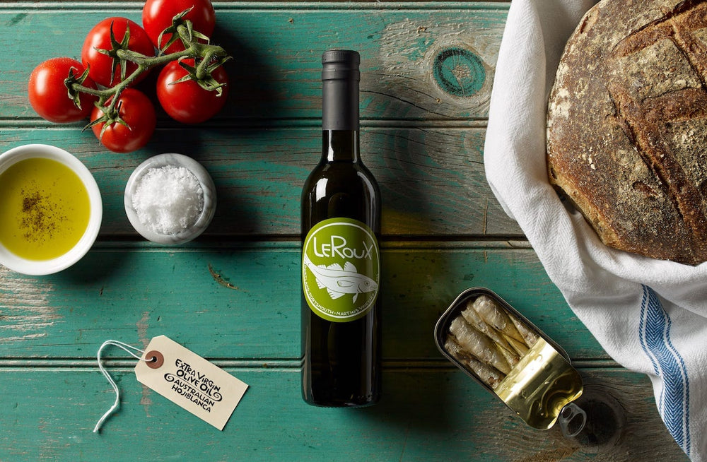 Olive oil bottle, fresh tomatoes, sea salt, artisan bread, and sardines on rustic wood table.