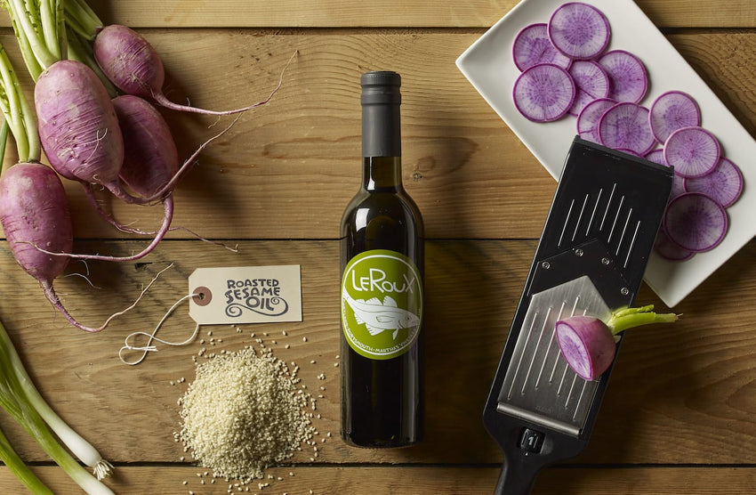 Sliced purple radishes on a plate, bottle of Leroux sesame oil, and raw sesame seeds.