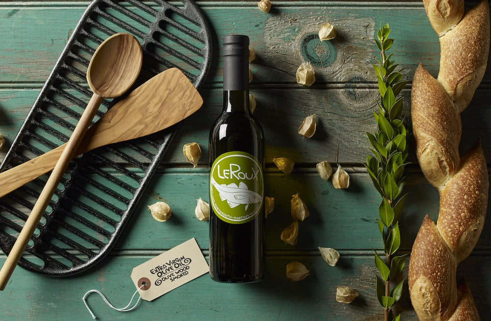 Olive oil bottle with label, fresh baguettes, wooden utensils, and grill pan on teal background.