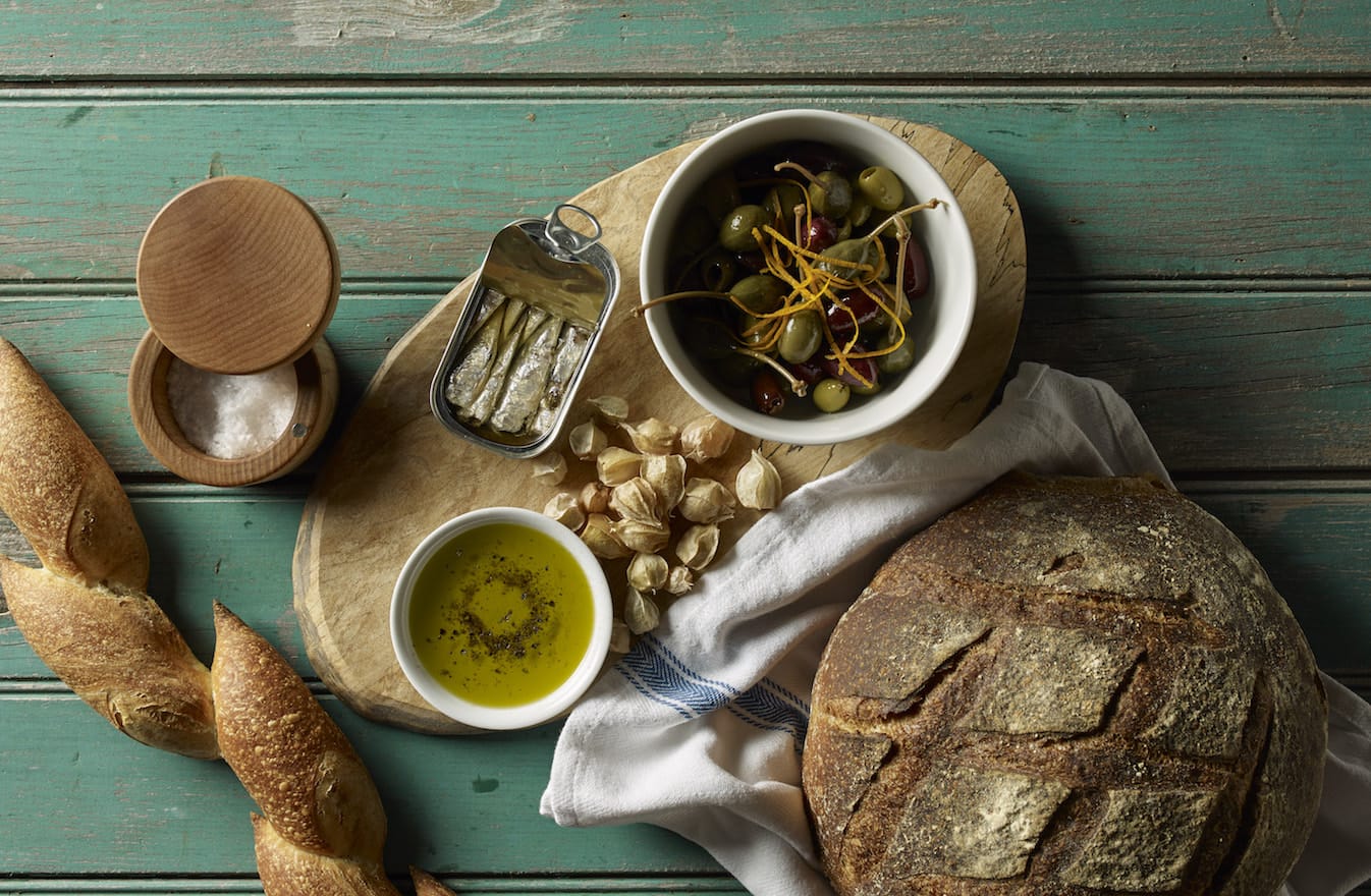 Rustic bread with olive oil, olives, sardines, and sea salt on worn wooden table.