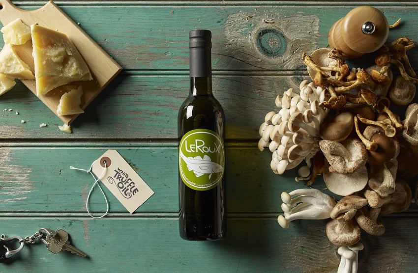 Bottle of Le Roux olive oil, assorted mushrooms, cheese wedge, and keys on wooden table.