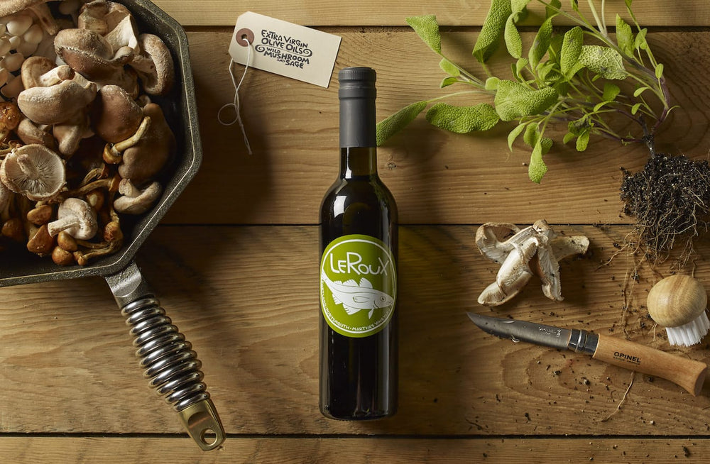 Variety of fresh mushrooms in a pan with olive oil bottle and herbs on wooden surface.
