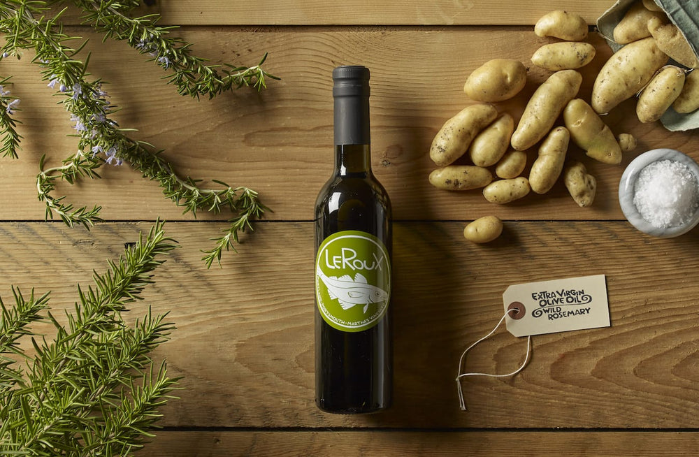 Extra virgin olive oil bottle with rosemary sprigs, potatoes, and salt on wooden table.