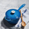 A Le Creuset Signautre Saucepan in Marseille blue sits on a marble countertop alongside a wooden spoon and dish towel.