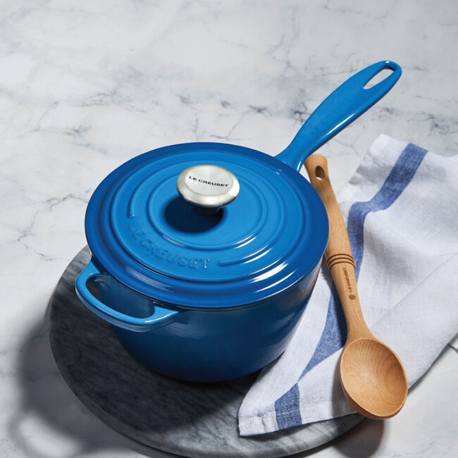 A Le Creuset Signautre Saucepan in Marseille blue sits on a marble countertop alongside a wooden spoon and dish towel.