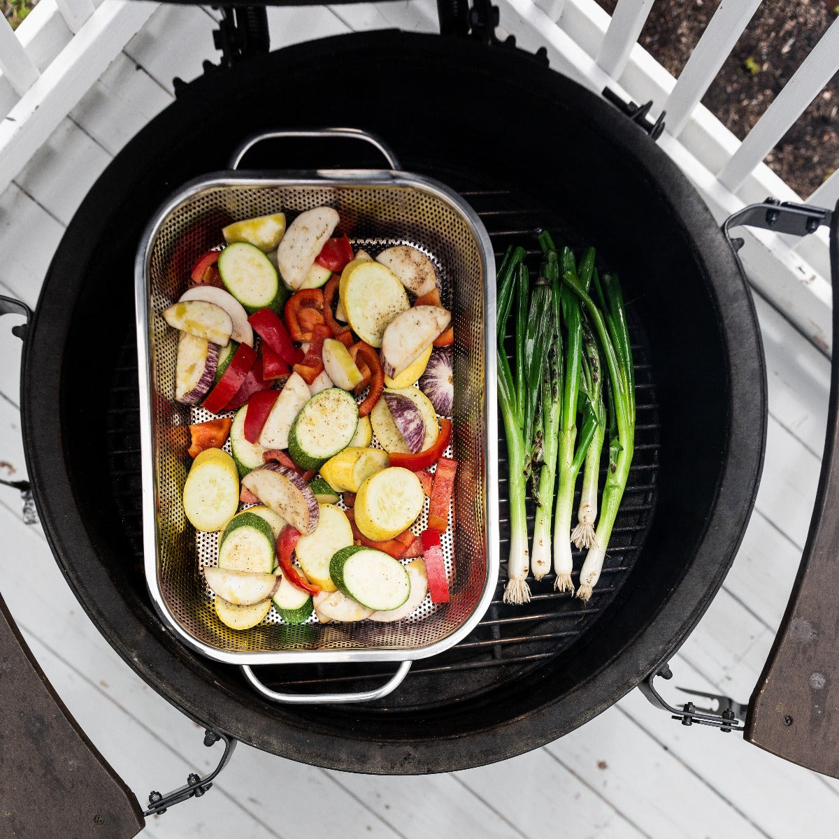 An RSVP Endurance Pierced Roasting Pan full of slices veggies sits on a grill, alongside roasting green onions.