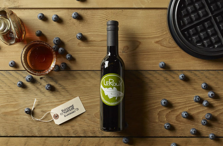 Bottle of LeRoux vinegar with blueberry tag, honey jar, fresh blueberries, and waffle iron on wooden table.